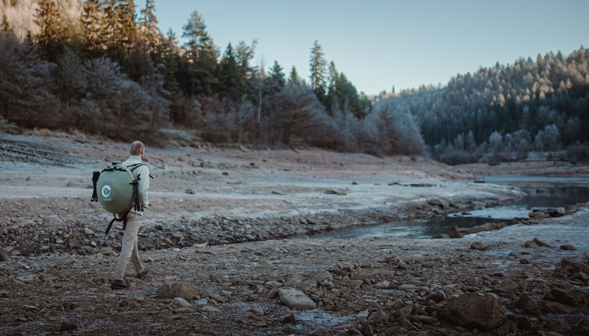 A person with a large green backpack walks across a rocky, dry riverbed surrounded by trees and hills under a clear sky—perhaps searching for inspiration after visiting Yatao Handpan Shop to buy handpan instruments.