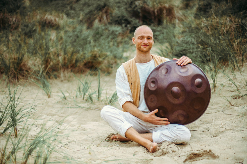 Malte Marten sits cross-legged on sandy ground with grass tufts, smiling and holding a brown handpan drum from Yatao Handpan Shop. He wears white clothes and a beige vest, surrounded by nature. Perfect for those looking to Handpan Kaufen.
